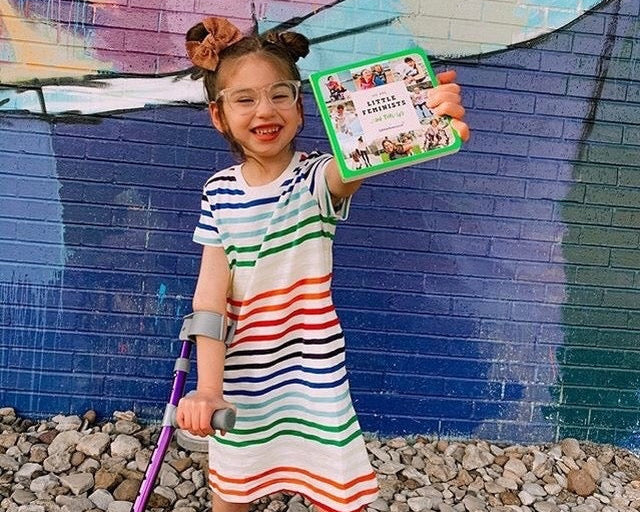 A smiling child with glasses and a forearm crutch holds up a Little Feminist book in front of a mural.