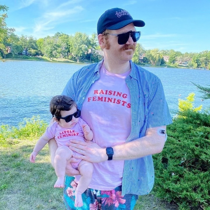 An adult and baby in matching pink feminist shirts beside a lake.